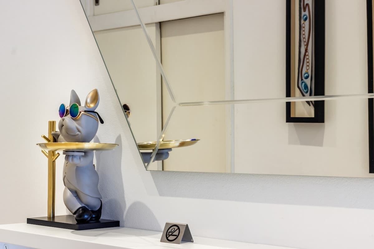 Escultura de um coelho com óculos sobre um pedestal, uma obra de arte emoldurada na parede e um aviso de proibido fumar no chão.