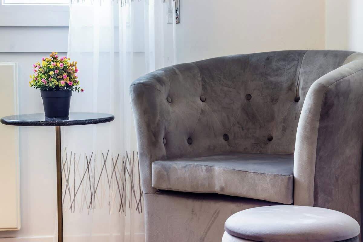 Un fauteuil à revêtement gris, doté d’un dossier capitonné et accompagné d’un pouf assorti, est placé à côté d’une petite table ronde sur laquelle se trouve un pot de fleurs.