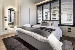 A modern bedroom with a large bed, a mirror, and a view of buildings through large windows. - [Lungolago] Nuovo Bilocale 3 (Como)