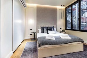 Ein modernes Schlafzimmer mit einem großen Bett, einem Nachttisch, einem Fenster mit Blick auf ein Geb äude und einer Klimaanlage.