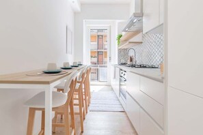 Et smalt køkken med et træbord og stole, hvide skabe og udsigt til en balkon.