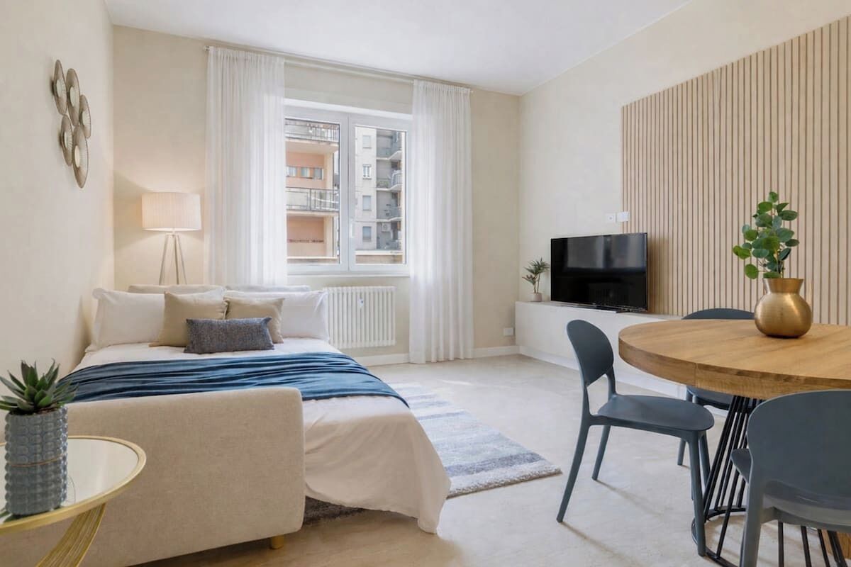 Une chambre d’hôtel avec un lit, une télévision, une table à manger avec des chaises, une fenêtre avec des rideaux et une plante.