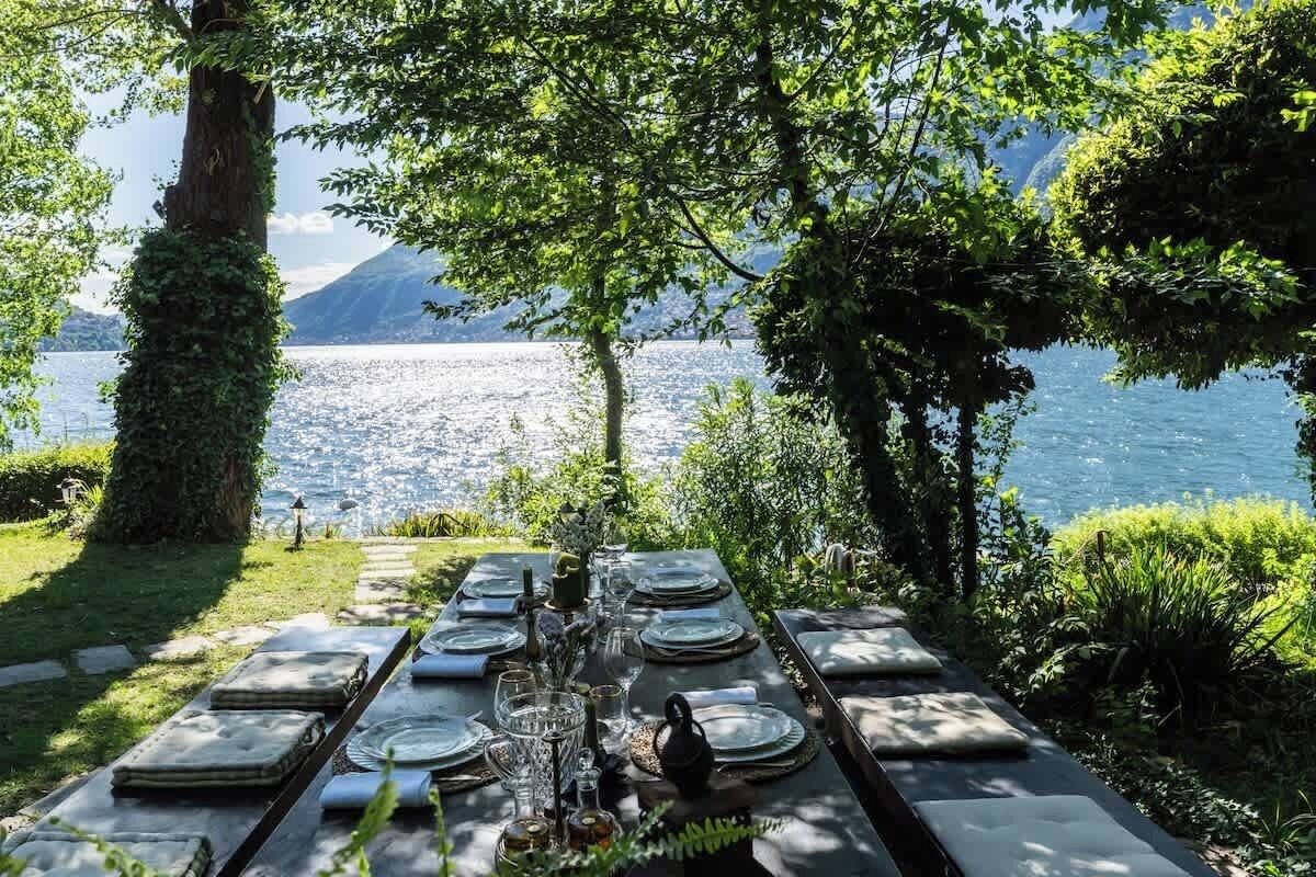 An outdoor dining table set up with plates, glasses, and cutlery, overlooking a body of water and mountains.