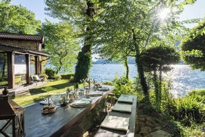 An outdoor dining area with a view of a lake, a pergola, and a stone table set for a meal. - Private Villa Pied en l'eau [Only by Boat] (Riva)