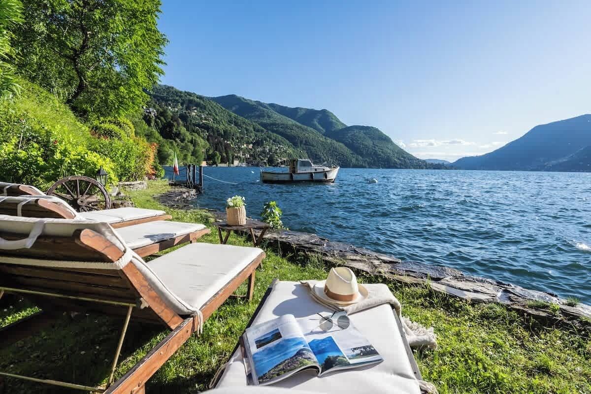 Un paisaje junto al lago con un bote, sillas de madera para relajarse y una mesa con un sombrero y una revista.