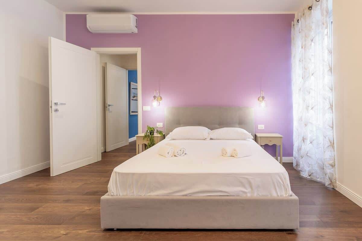 Une chambre avec un grand lit, une table de chevet, une commode, un climatiseur fixé au mur et une porte donnant sur une autre pièce.