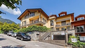 A multi-story residential building with a gated entrance, parked cars, and a mountainous backdrop.