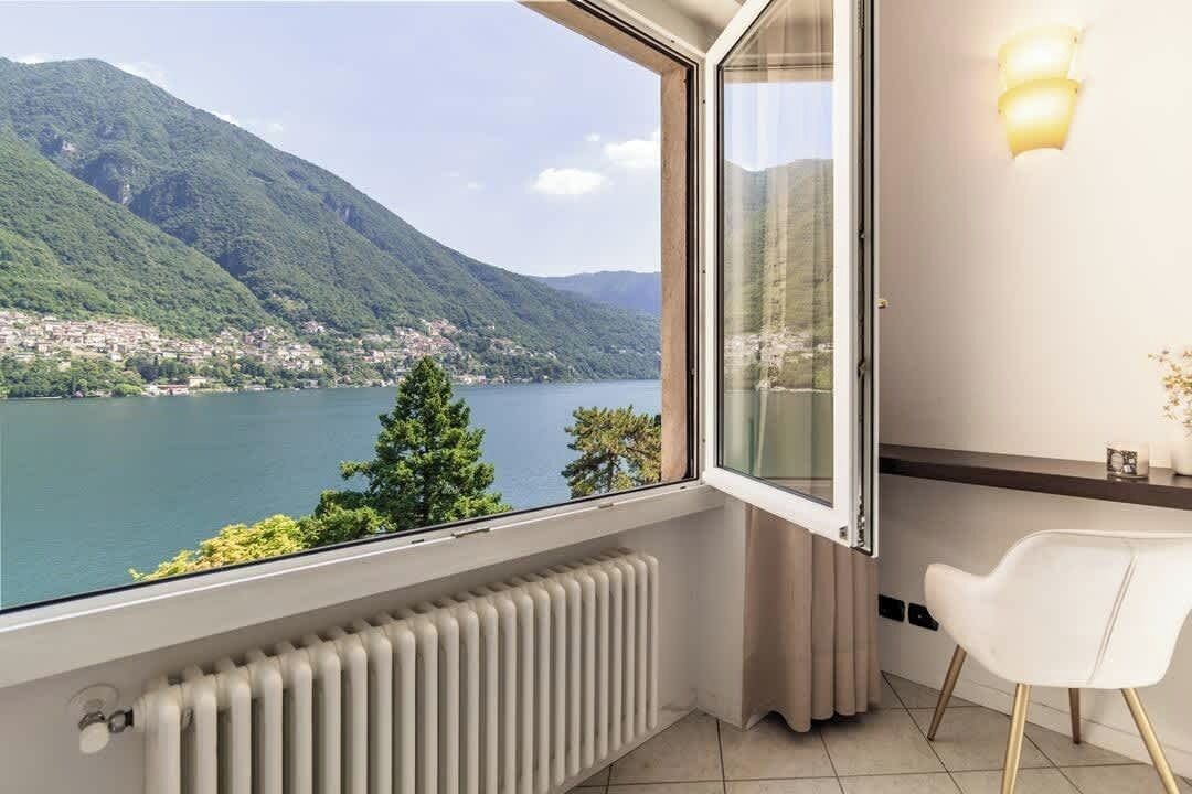 A room with a view of a lake and mountains, a white radiator, a white chair, and a wall-mounted light fixture.