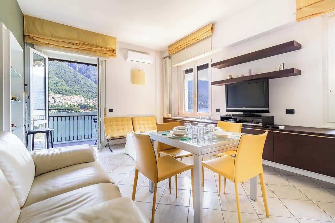 A modern living room with a sofa, dining table, and chairs, a kitchen area with a TV, and a balcony with a mountain view.