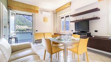 A modern living room with a sofa, dining table, and chairs, a kitchen area with a TV, and a balcony with a mountain view.