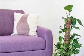 Ein violettes Sofa mit einem weiß-violetten Kissen und einer hohen Pflanze mit grünen Blättern.