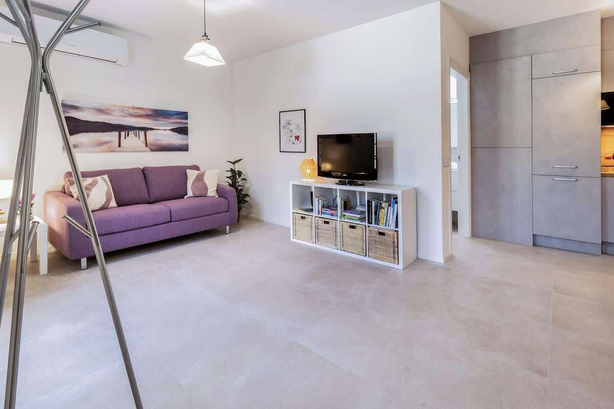 A modern living room with a sofa, a television on a white stand, and a kitchen area with cabinets.