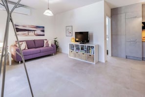 Ein modernes Wohnzimmer mit einer Couch, einem Fernseher auf einem weißen Schrank und einer Küchenzeile mit Schränken.