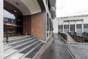 A building with a staircase leading to a covered entrance, a parking area, and a sign that reads 'STADIO G.SINIGAGLIA'. - [Lungolago] Nuovo Bilocale 2 (Como)