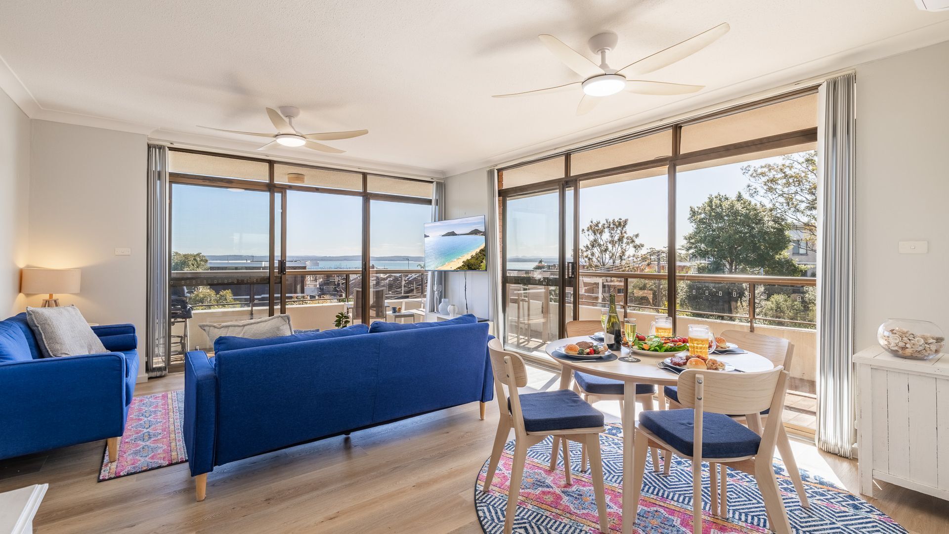 A modern living room with a blue sofa, a dining table with chairs, and large windows offering a view of the sea.