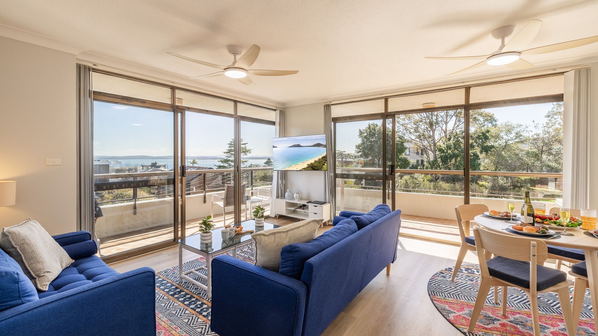 Ein modernes Wohnzimmer mit einer Couch, einem Esstisch mit Stühlen und einem an der Wand befestigten Fernseher. Es gibt einen Balkon mit Meerblick.
