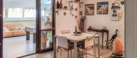A dining area with a white table, chairs, and a glass partition. There are decorative items on the walls and a view of a living room with a sofa and a window.