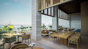 An outdoor dining area with tables and chairs, a view of the ocean, and tropical plants.