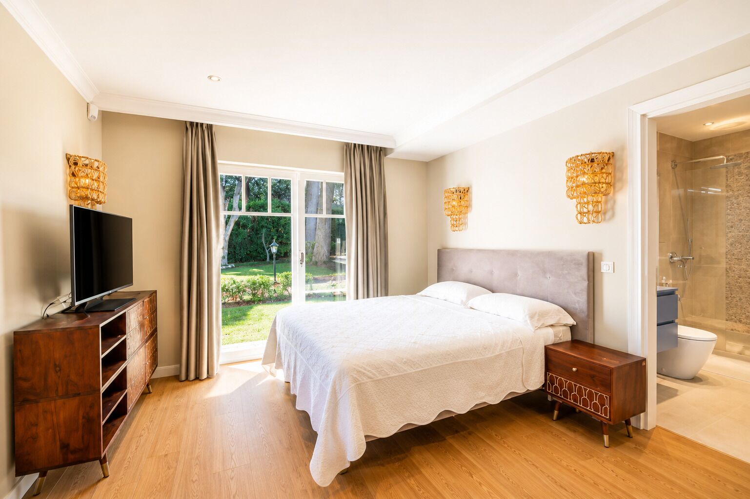 A bedroom with a bed, a TV, a wooden dresser, a bathroom with a shower, and a view of a garden.