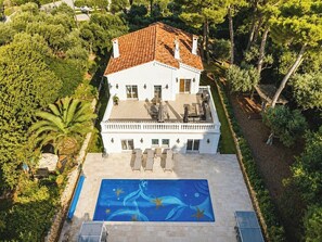A white building with a tiled roof, a swimming pool with a blue mural, and a garden with various trees. - Five-bedroom villa on Cap d'Antibes (Juan les Pins Antibes)