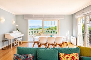 Ein modernes Wohnzimmer mit einem Klavier, einem Essbereich mit Tisch und Stühlen, einem grünen Sofa und Blick ins Grüne durch große Fenster.