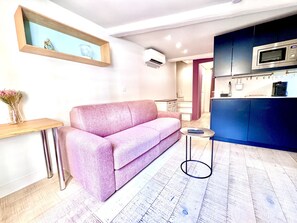 A modern living space with a pink sofa, a small round table, a wooden side table, and a kitchen area with a microwave. - Cocon Bonaparte 2 - Garibaldi (Nice)