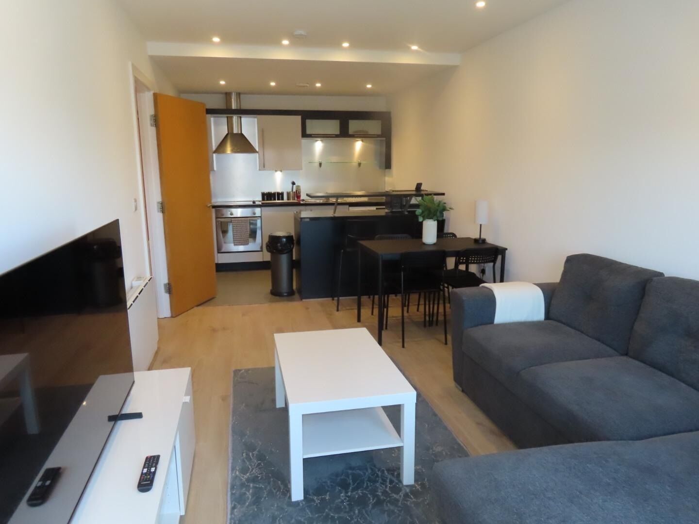 Ein modernes Wohnzimmer mit einem grauen Sofa, einem weißen Couchtisch und einem Fernseher. Im Hintergrund ist eine Küche mit Essbereich zu sehen.