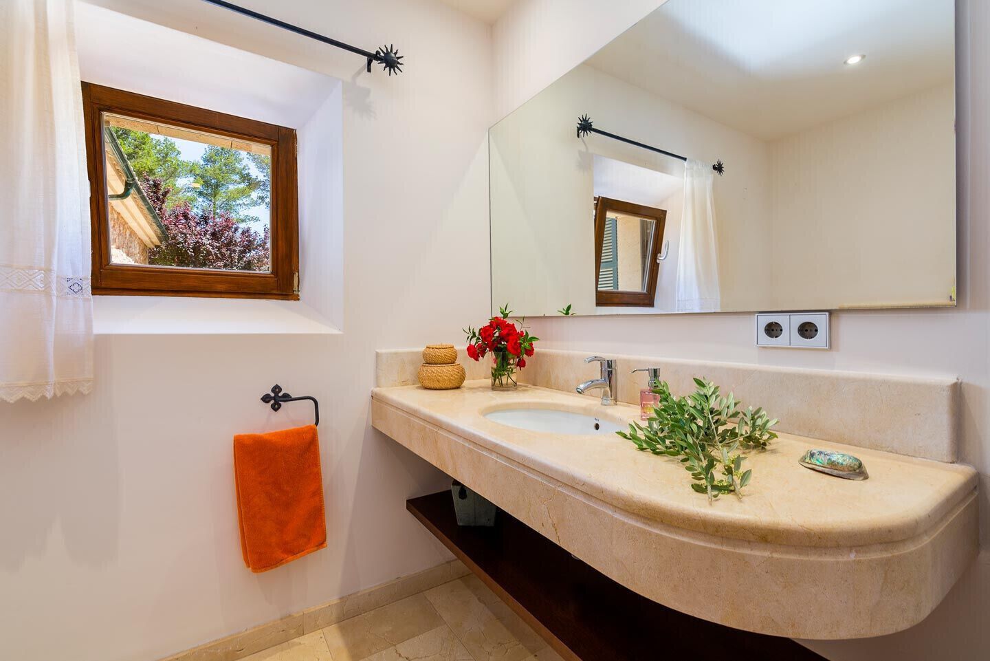 A bathroom with a double sink, a large mirror, a window with a view of trees, and a towel rack with an orange towel.