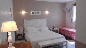 Un dormitorio con cama, mesitas de noche, lámpara, banco, ventilador de pared y ventana con cortinas.