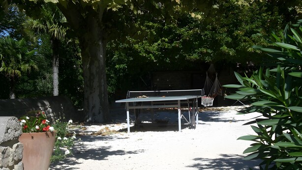 Un área de estar al aire libre cubierta, con una mesa y sillas, rodeada de árboles y un muro de piedra.