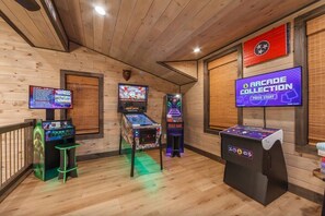 Een kamer met houten wanden en plafond, met flipperspelautomaten, een groot televisiescherm en een houten bank.