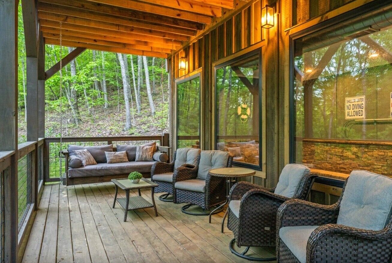 Un espacio al aire libre techado con sofá y sillas, una mesita y vista al bosque.