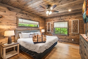 A wooden cabin bedroom with a bed, nightstand, lamp, ceiling fan, and windows with a view. - UndHrvstMoon: Pool, HotTub, Wifi, Theater, Games, Pets, OutdoorFireplace (Sevierville)