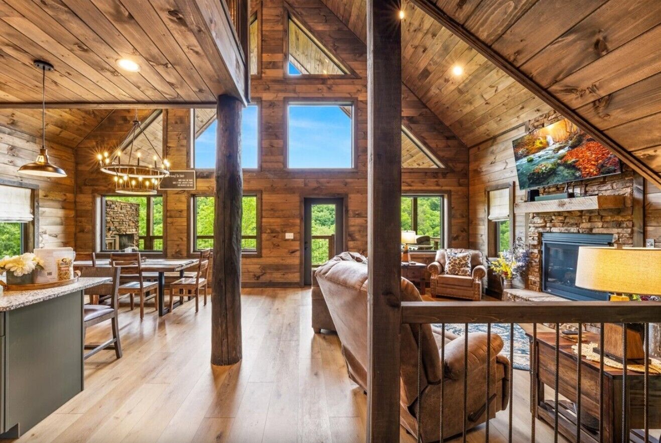 A spacious log cabin interior with a dining area, a living room with a fireplace, and a large window overlooking greenery.