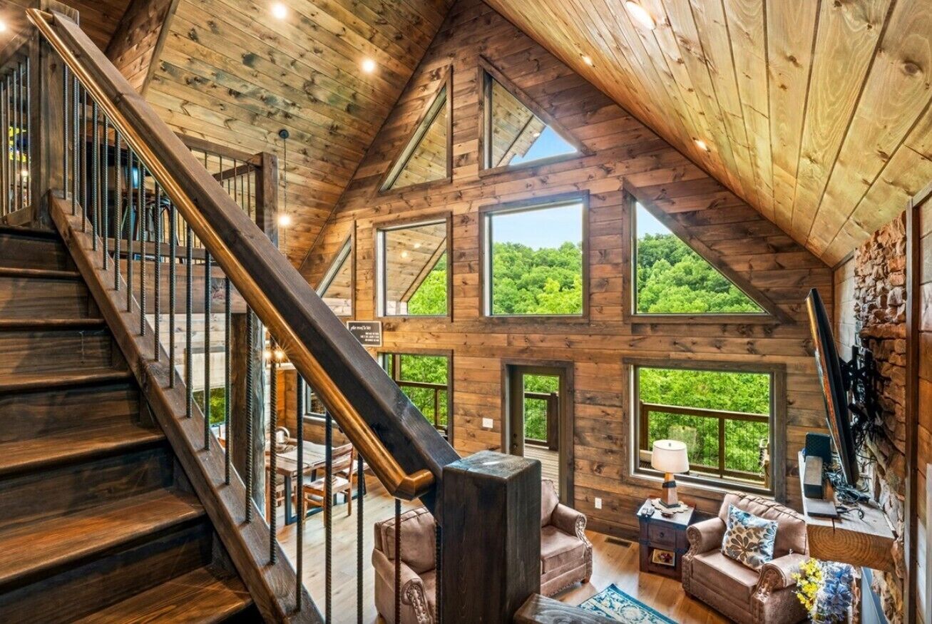 A spacious loft with a wooden staircase, large windows offering a view of greenery, and a cozy seating area with a lamp.