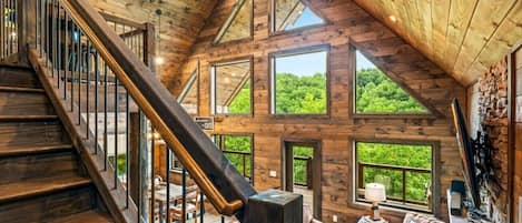 Un loft espacioso con una escalera de madera, ventanas amplias que dan a vistas de vegetación y una zona de estar acogedora con una lámpara.