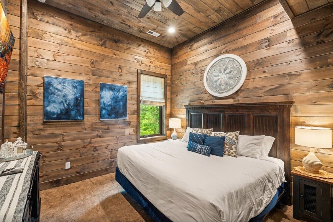 A wooden-paneled bedroom with a large bed, two wall-mounted paintings, a round decorative item above the bed, and a window with a view of greenery.