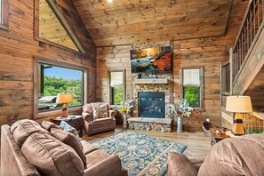 A log cabin interior with a fireplace, wooden walls, and a large window overlooking greenery. - UndHrvstMoon: Pool, HotTub, Wifi, Theater, Games, Pets, OutdoorFireplace (Sevierville)
