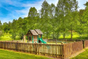 A wooden playhouse with a slide, surrounded by a wooden fence, set in a grassy park with trees. - PeacefulCreek: Pool, HotTub, Wifi, FirePit, Pets, Creek, Resort Facilities (Sevierville)
