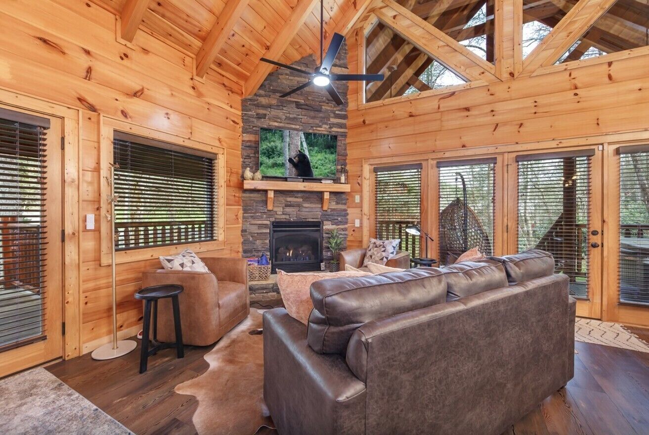A log cabin interior with a stone fireplace, leather sofas, and a ceiling fan.