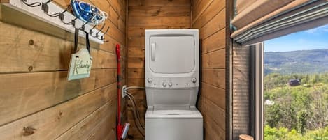 A compact room with a washing machine, a drying rack, and a window with a view of the outdoors.