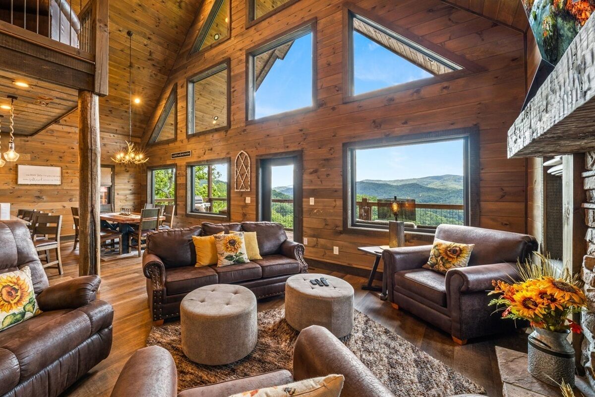 A spacious living room with leather sofas, ottomans, and a fireplace. Large windows offer views of the outdoors.