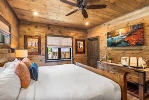 A wooden cabin bedroom with a bed, a ceiling fan, a window, and a painting on the wall. - SerenityLod: POOL, Theater, Games, HotTub, Wi-Fi, Pets (Sevierville)