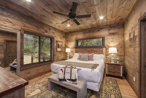 Une chambre à coucher aux murs lambrissés, comprenant un lit, deux tables de chevet, un ventilateur de plafond et une fenêtre avec des stores.