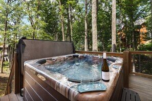 Un bain à remous avec une bouteille de vin et des verres, installé sur une terrasse entourée d’arbres.