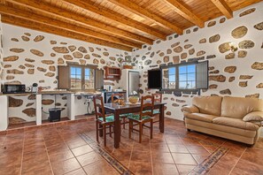 A room with a stone wall, wooden ceiling beams, a dining table with chairs, a sofa, and a television. - House 'Casa Clarita Tindaya' with Historic Charm and Views (Tindaya)
