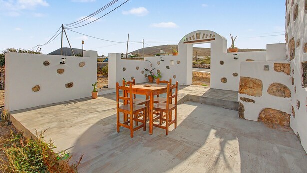 A patio with a wooden table and chairs, a white wall with stone accents, and a gate leading to a property. - Casa Clarita: House with Mountain Views, Private Terrace, and Wi-Fi (Tindaya)