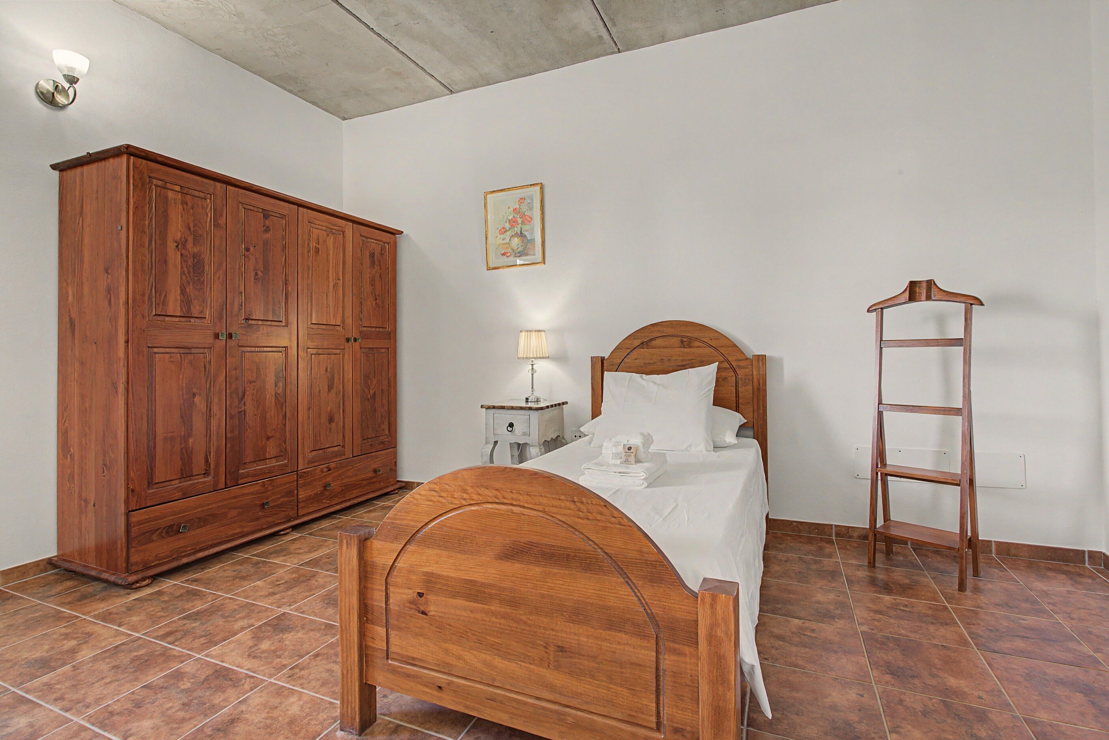 A bedroom with a wooden bed, a nightstand, a lamp, a wardrobe, and a painting on the wall.