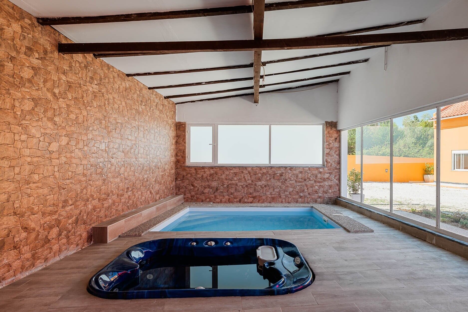 An indoor swimming pool with a hot tub, stone walls, and large windows.