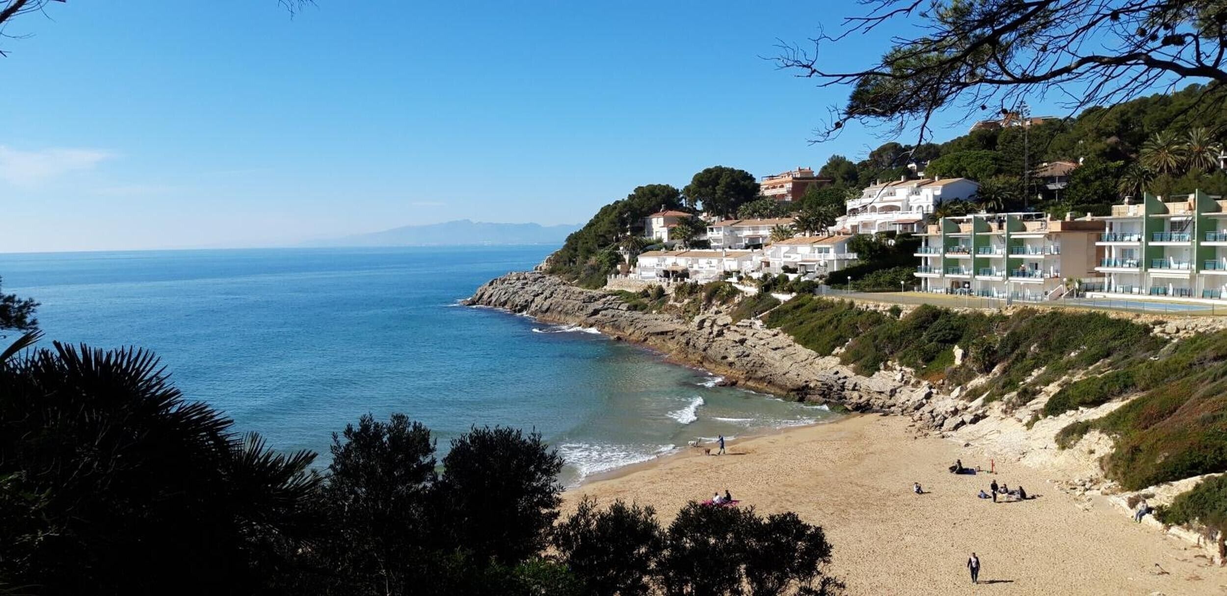 A coastal view with a beach, rocky shoreline, and buildings with balconies overlooking the sea.